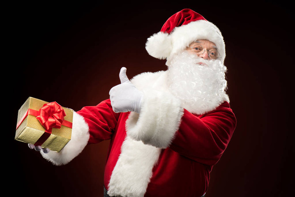Happy Santa Claus with gift box in hand showing thumb up - Фото, зображення
