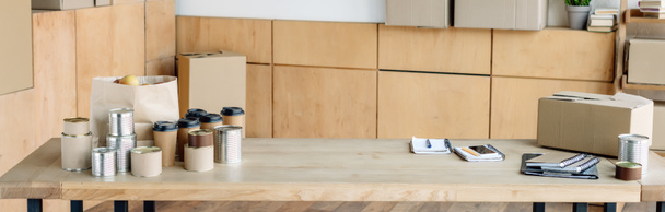 πανοραμική βολή από ξύλινο τραπέζι με κονσέρβες, φλιτζάνια μίας χρήσης, χάρτινη σακούλα και χάρτινο κουτί  - Φωτογραφία, εικόνα