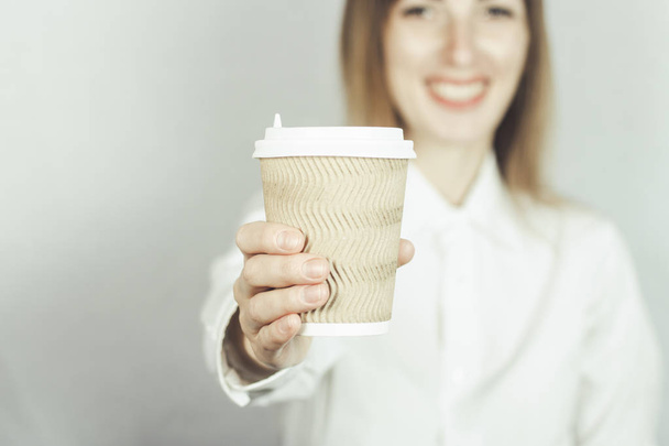 Ένα κορίτσι με άσπρη μπλούζα της κρατά το χέρι της μπροστά σε ένα χάρτινο φλιτζάνι καφέ ή τσαγιού. Ιδέα φαγητού σε πακέτο, καφετέρια. - Φωτογραφία, εικόνα