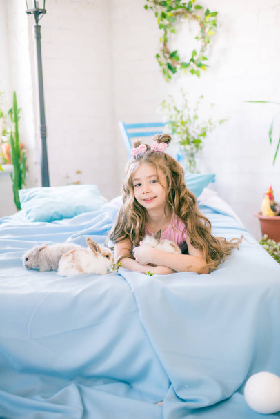 Little cute girl with long curly hair on the bed with little bunnies and Easter decor at home. - Foto, afbeelding