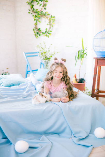 Little cute girl with long curly hair on the bed with little bunnies and Easter decor at home. - Photo, Image