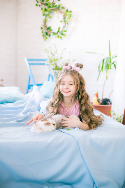 Little cute girl with long curly hair on the bed with little bunnies and Easter decor at home. - Zdjęcie, obraz