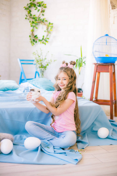 Klein schattig meisje met lange krullend haar op het bed met kleine konijntjes en Pasen decor thuis. - Foto, afbeelding