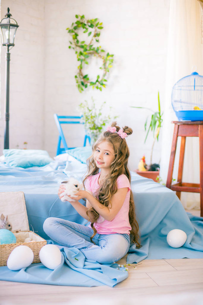 Little cute girl with long curly hair on the bed with little bunnies and Easter decor at home. - Fotografie, Obrázek