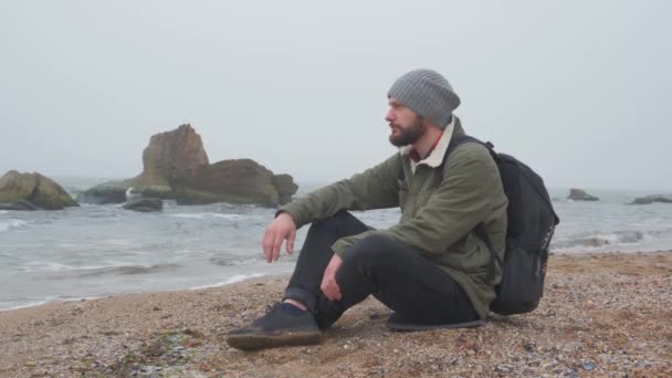 Mladý vousatý muž sedí večer sám na pláži a hází kamínky do vody - Záběry, video