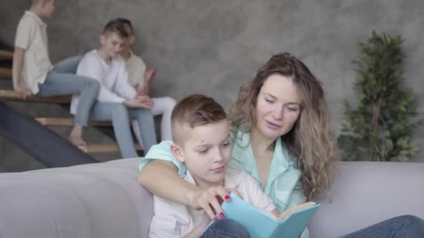 Portrait of pretty young mother reading book to her son while the rest of her teen kids playing with each other in the background sitting on stairs with tablet. Big friendly noizy family - Video