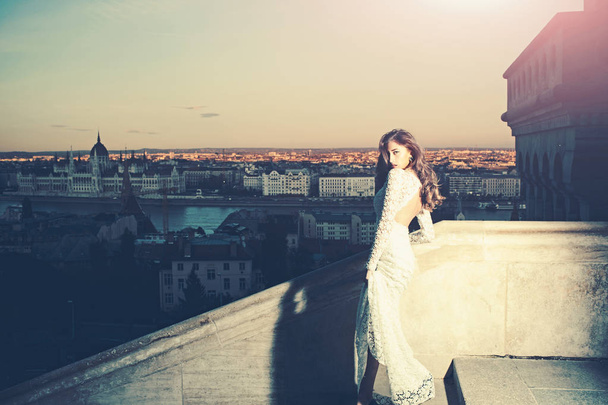 Woman in white wedding dress on evening city view, fashion. Sensual woman with long hair on balcony, beauty. Bride with makeup. Girl with glamour look in wedding - Photo, Image