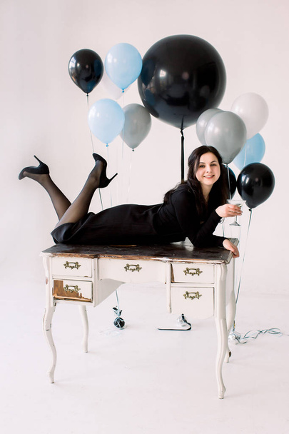 Attractive young girl in black dress lying on white vintage table, holding the martini glass, on white background. Pretty girl elebrating Happy New Year, birthday party balloons on the background - Fotó, kép