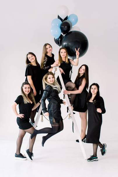 Happy young seven girls in black dresses having fun celebrating birthday in light room. The friends having fun posing on the ladder, decorated with colorful air balloons. - Fotografie, Obrázek