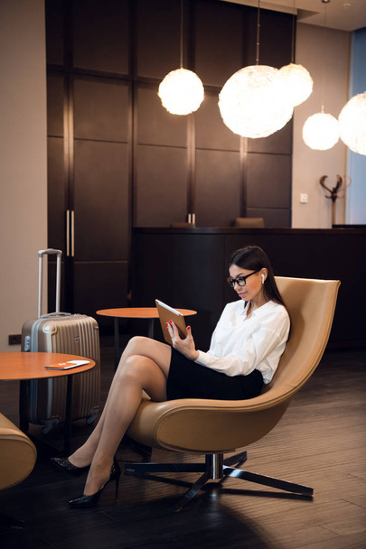 Уверенная деловая женщина слушает музыку на планшетном компьютере, сидя в кресле в бизнес-зале аэропорта - Фото, изображение