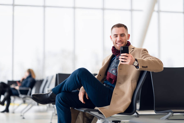 Atrakcyjny młody mężczyzna w stylowym szaliku i płaszczu, siedzący wewnątrz terminalu na lotnisku. Patrzy na ekran telefonu komórkowego i uśmiecha się - Zdjęcie, obraz