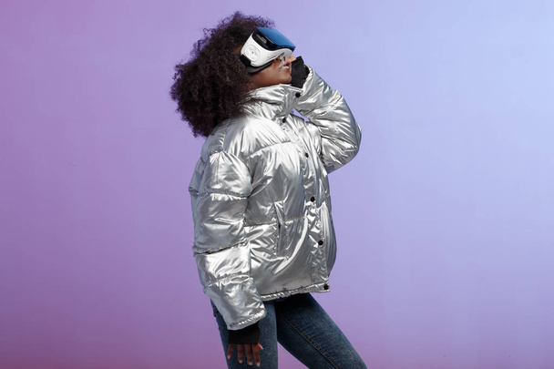 Mod curly brown-haired girl dressed in a silver-colored jacket uses the virtual reality glasses in the studio on neon background - Foto, imagen