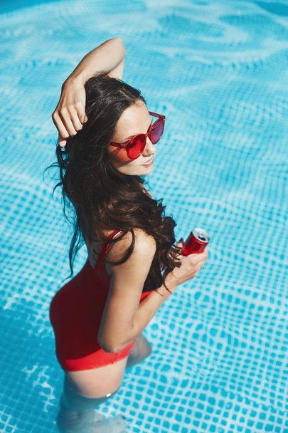 Fashionable and happy brunette model woman with a perfect sexy figure in stylish red swimsuit and trendy red sunglasses in the swimming pool holding a cold drink in her hand - Φωτογραφία, εικόνα