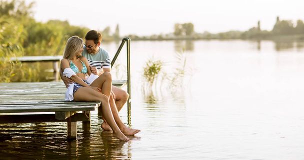 Loving couple sitting on the pier on lake at summer sunset - Photo, Image