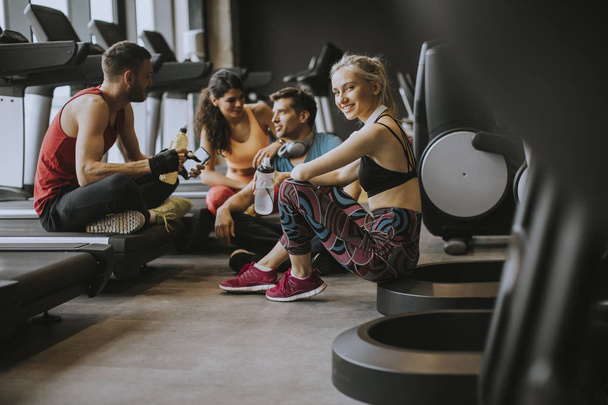 Группа молодых людей в спортивной одежде разговаривают и смеются вместе, сидя на полу спортзала после тренировки
 - Фото, изображение