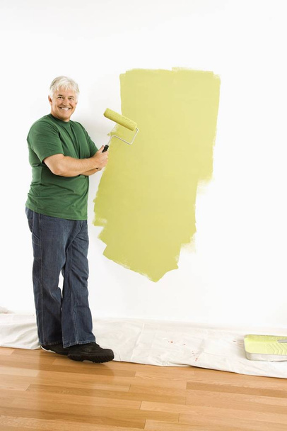 Middle-aged man painting wall green with paint roller smiling at viewer. - Foto, Imagen