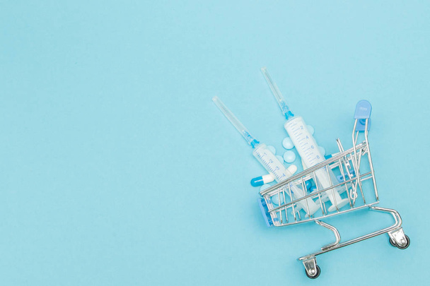 Pills and medical injection in shopping trolley on blue background. Creative idea for health care cost, drugstore, health insurance and pharmaceutical company business concept. Copy space - Foto, afbeelding