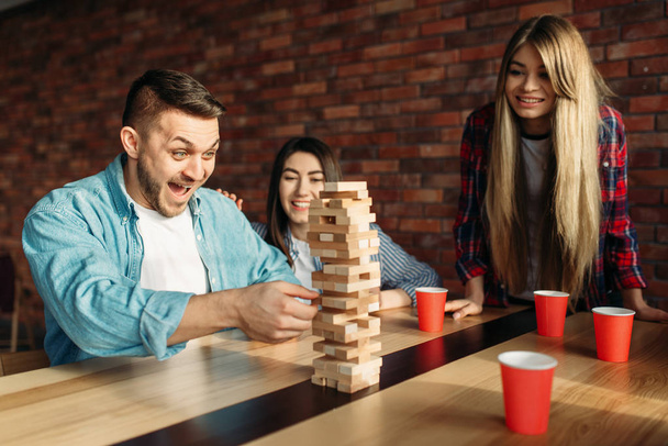 Smiling friends plays table game at home, selective focus on tower. Board game with wooden blocks requiring high concentration, entertainment for funny company - Foto, Bild