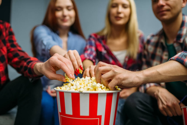 Group of people eating popcorn and having fun in cinema hall before the screening. Male and female youth sitting on sofa in movie theater - Foto, imagen