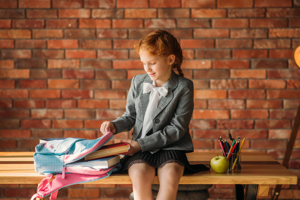 Милая школьница кладет учебник в школьную сумку, вид сбоку. Женщина-ученица с рюкзаком, стол с учебниками, яблоко и глобус на backgrpund, молодая девушка в школе
 - Фото, изображение