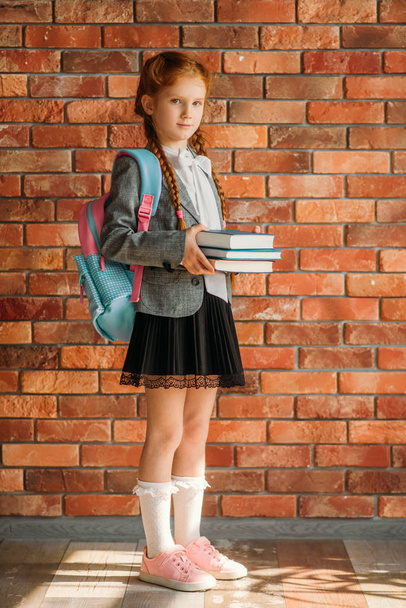 Cute schoolgirl with schoolbag holds books, brick wall on background. Adorable female pupil with backpack and books poses in the school - Foto, Bild
