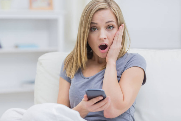 Portrait of casual shocked young woman with cellphone on sofa at home - Фото, изображение