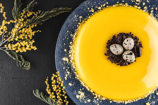 Пасхальный торт с желтым зеркалом глазури, шоколад, весенние цветы, перепелиные яйца на темном каменном фоне. С праздником Пасхи. Вид сверху, плоский
 - Фото, изображение