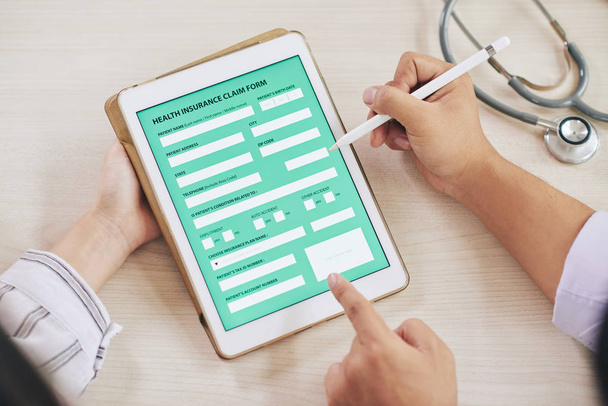 Médico con estilo apuntando a la pantalla táctil con formulario digital de reclamo de seguro de salud mientras lo discute con el paciente
 - Foto, imagen