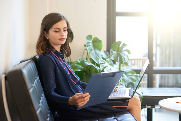 Bella giovane donna d'affari lettura relazione finanziaria allegata agli appunti quando si lavora su laptop in ufficio
 - Foto, immagini