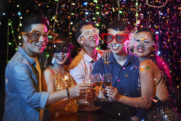 Happy excited young Vietnamese people in party glasses posing with champagne glasses under falling confetti - Photo, Image