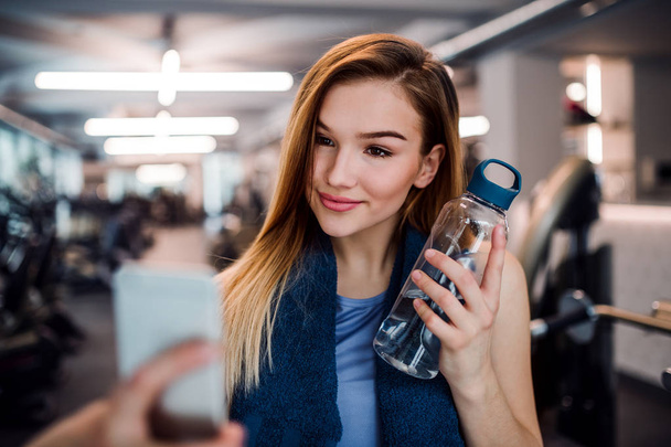 A portrait of young girl or woman with smartphone in a gym, taking selfie. - Foto, Bild