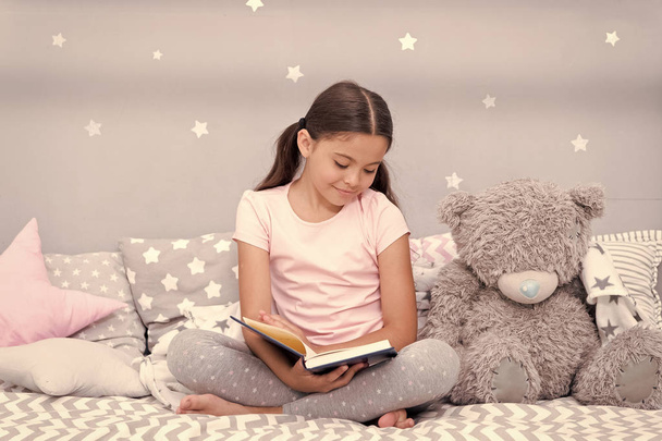Read before sleep. Girl child sit bed with teddy bear read book. Kid prepare to go to bed. Pleasant time in cozy bedroom. Girl kid long hair cute pajamas relax and read book to bear toy - 写真・画像