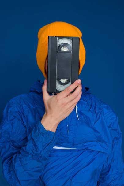 young man  in 90s sports jacket and yellow hat with VHS cassette on blue background - Fotó, kép