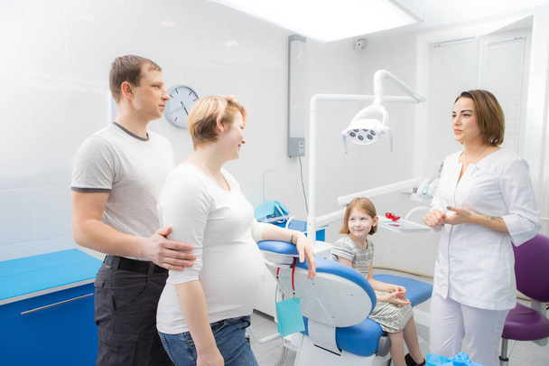 Een vrouwelijke tandarts communiceert met een kind en haar familie in een tandheelkundige kliniek. De moeder van het meisje is zwanger - Foto, afbeelding