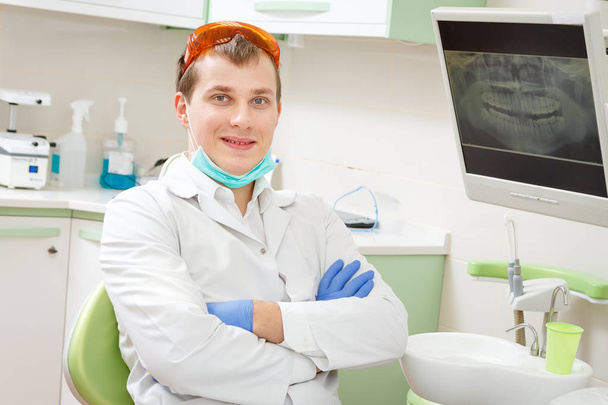 Πορτρέτο του νεαρού χαμογελαστή οδοντίατρο που κάθονται στη σύγχρονη οδοντιατρική χειρουργική επέμβαση στο οδοντιατρείο - Φωτογραφία, εικόνα