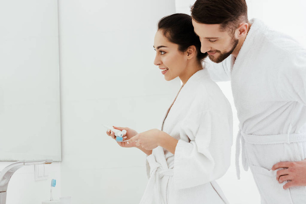 Veselý vousatý muž s pohledem na šťastnou ženu, držící zubní kartáček a zubní pastu  - Fotografie, Obrázek