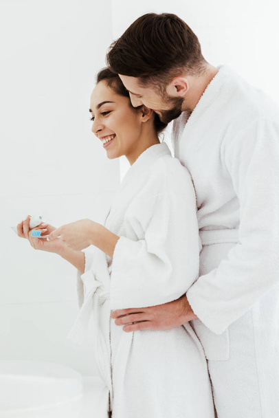 happy man hugging cheerful girlfriend holding toothbrush and toothpaste  - Foto, Imagen