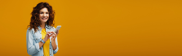 plano panorámico de atractiva pelirroja rizada sosteniendo teléfono inteligente en naranja
  - Foto, imagen