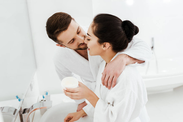 cheerful boyfriend kissing happy girl holding container with face cream  - Foto, Bild