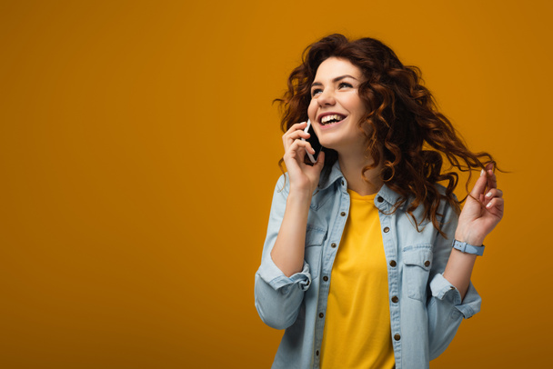 šťastná kudrnatá žena, která mluví po telefonu a dotýká se vlasů na oranžovém  - Fotografie, Obrázek
