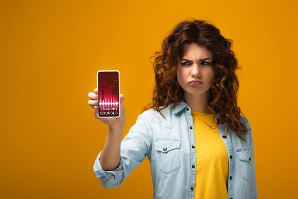 upset redhead woman holding smartphone with trading courses on screen on orange  - Fotografie, Obrázek