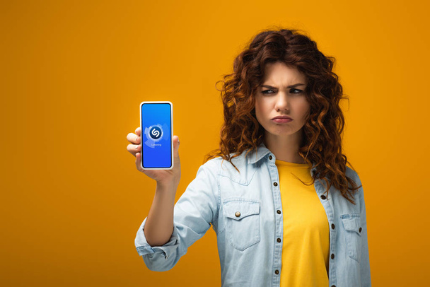 upset redhead woman holding smartphone with shazam app on screen on orange  - Zdjęcie, obraz