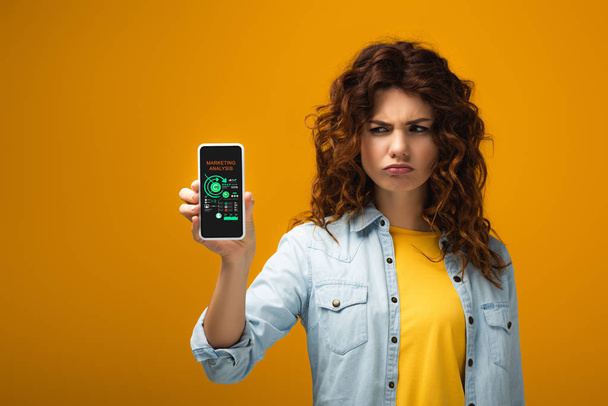 upset redhead woman holding smartphone with marketing analysis on screen on orange  - Fotografie, Obrázek