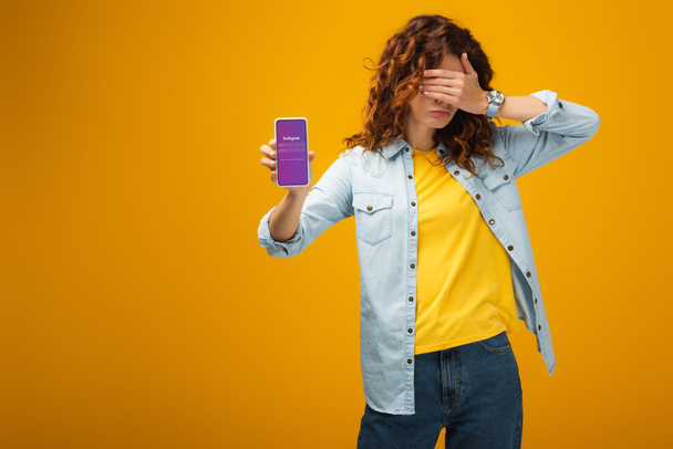 redhead woman covering eyes and holding smartphone with instagram app on screen on orange  - Foto, afbeelding