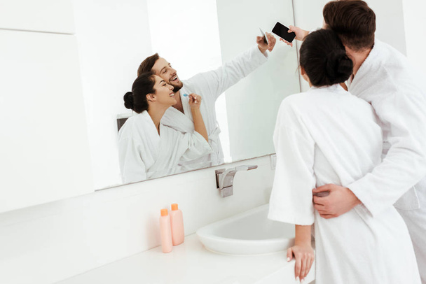 vrolijke man knuffelen gelukkige vrouw in badjas terwijl het nemen van selfie in de badkamer  - Foto, afbeelding