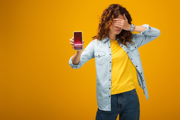 rothaarige Frau verdeckt Augen und hält Smartphone mit Handelskursen auf dem Bildschirm auf orange  - Foto, Bild