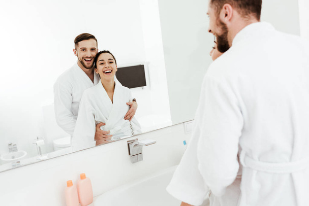 selective focus of bearded man looking at mirror with cheerful brunette woman while hugging in bathroom  - Foto, Imagem