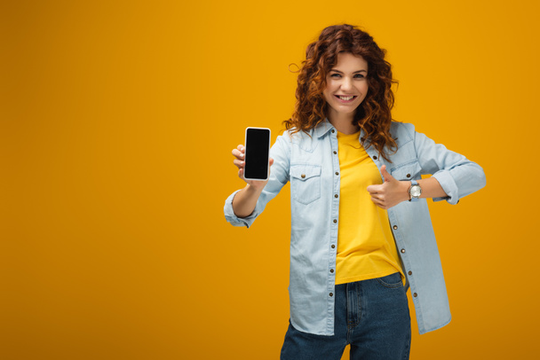 mujer pelirroja feliz sosteniendo teléfono inteligente con pantalla en blanco y mostrando el pulgar hacia arriba en naranja
  - Foto, imagen