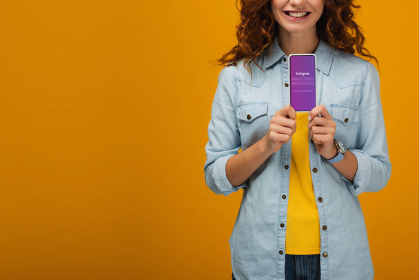 cropped view of cheerful curly woman holding smartphone with instagram app on screen on orange  - Foto, imagen