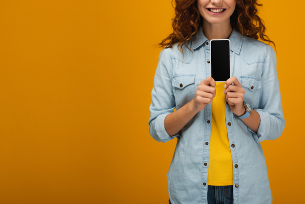 cropped view of cheerful curly woman holding smartphone with blank screen on orange  - Valokuva, kuva